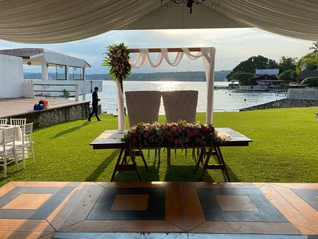 Montaje de boda frente al lago, con mesa de madera, flores y sillas bajo un techo de tela